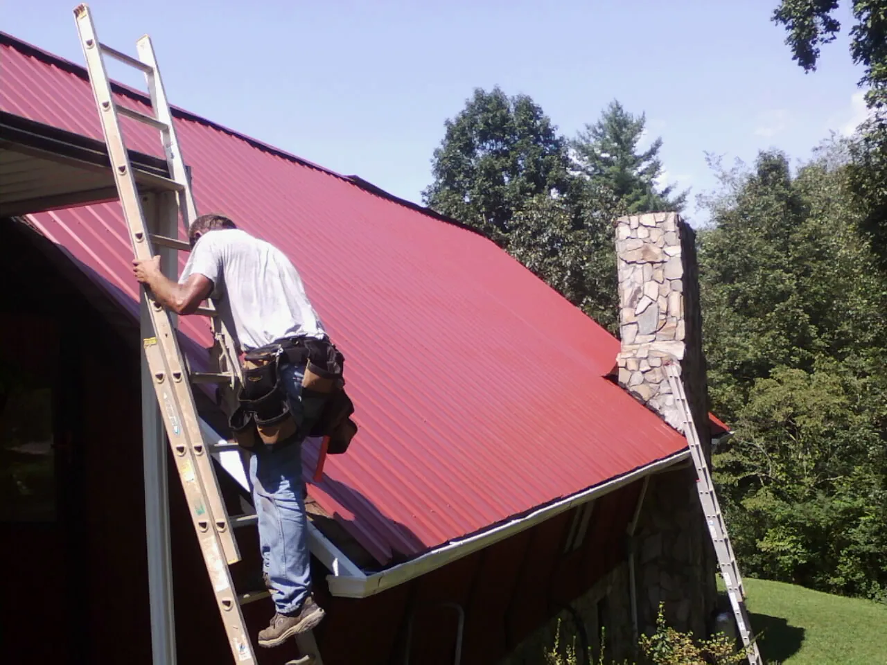 Roofer on a ladder beside a red metal roof and stone chimney — Soffit & Fascia Repair by Roofing Brothers in East Liberty